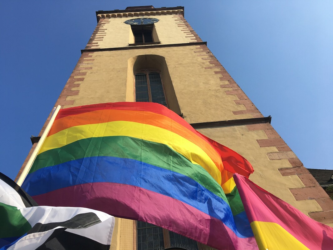 Froschperspektive des Kirchturms der Katharinenkirche in Frankfurt, vor der unten eine Regenbogenflagge weht.